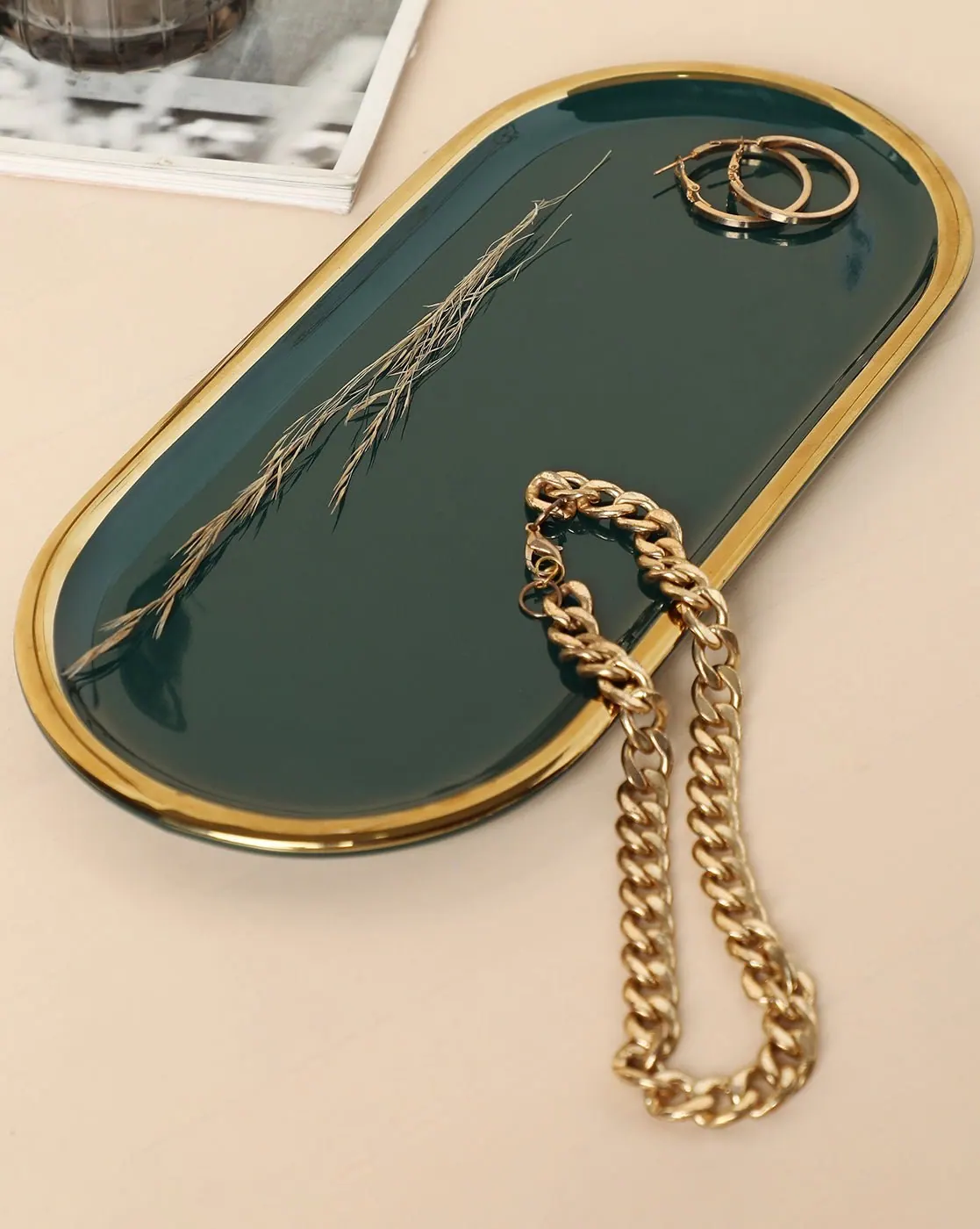 An oval forest-green ceramic tray with a gold rim holding jewelry